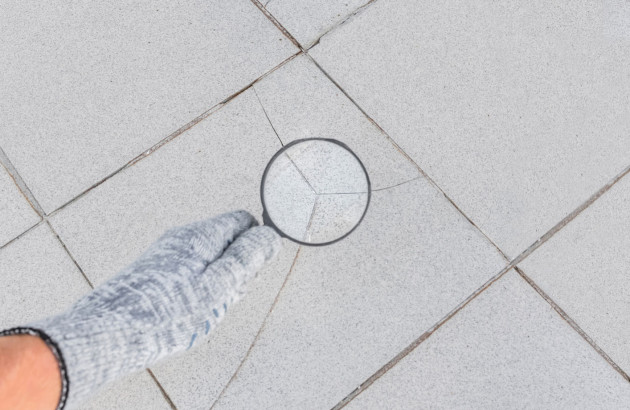 Carrelage gris fissuré, une main gantée montre la fissure à l'aide d'une loupe