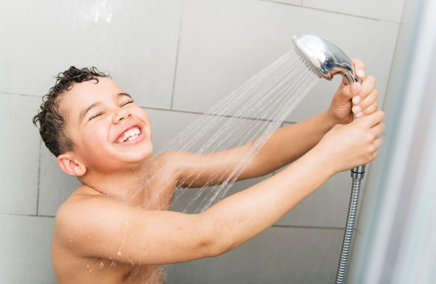 enfant souriant qui prend une douche et tient le pommeau avec l'eau qui s'écoule sur un fond de carrelage gris