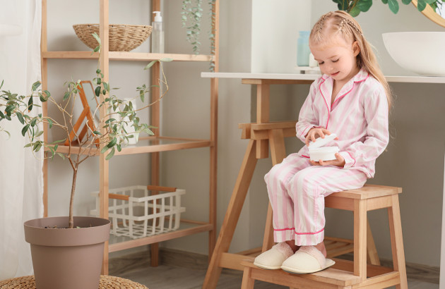 Petite fille en pyjama assise sur un marchepied en bois dans une salle de bains aux tons naturels.