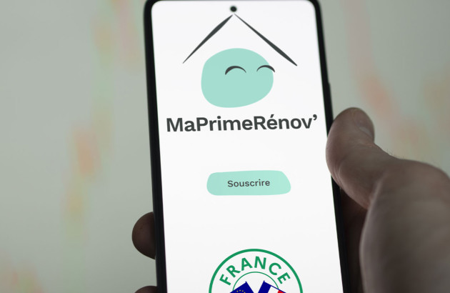 Un utilisateur tient un smartphone affichant l’application MaPrimeRénov’ avec le bouton “Souscrire” 