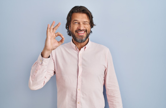 Homme en chemise rose devant un fond bleu qui fait un signe OK avec sa main
