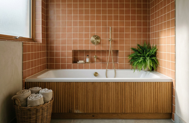 Baignoire rectangulaire avec habillage en bois, carrelage terracotta et robinetterie murale finition dorée.