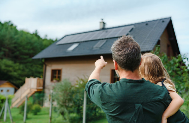 Homme portant une enfant dans ses bras et montrant les panneaux solaires installés sur le toit de leur maison.