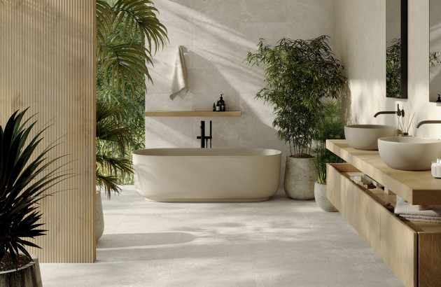 Salle de bains naturelle et lumineuse avec carrelage effet pierre beige, baignoire îlot et meuble double vasque en bois clair.