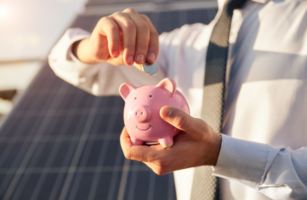 Homme en chemise devant une installation de panneaux solaires, il insère une pièce dans une tirelire cochon