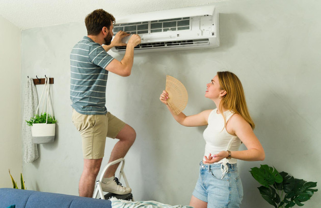 homme sur une petite échelle inspecte un climatiseur mural blanc et une femme se rafraîchit avec un éventail
