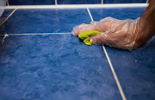 Nettoyage des joints sur carrelage bleu, main protégée essuyant les lignes de jointoiement avec une microfibre.