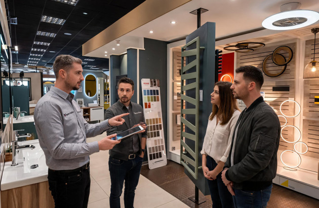 Conseiller présentant un radiateur sèche-serviettes à un couple dans un showroom spécialisé en équipements de chauffage et de salle de bains.