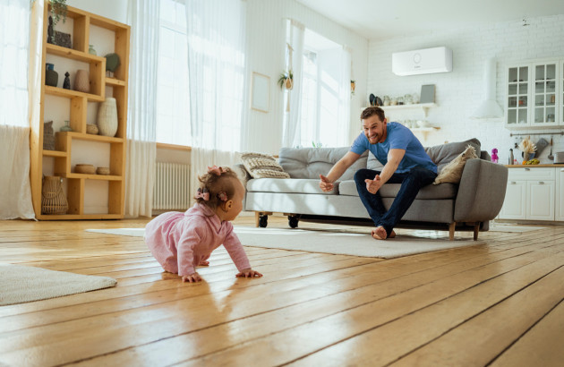  bébé qui rampe sur un parquet en bois vers un homme assis sur un canapé dans un salon lumineux avec un climatiseur mural 