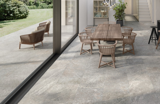 Salle à manger ouverte sur l’extérieur avec carrelage effet béton ciré, table en bois, chaises design et grandes baies vitrées.