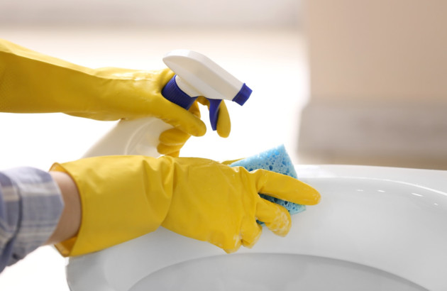Personne qui utilise des gants, du produit et une éponge pour nettoyer des toilettes