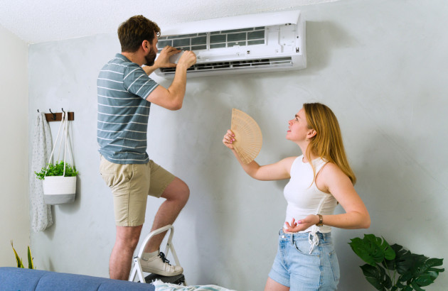 Technicien réparant une climatisation pendant qu’une cliente se rafraîchit avec un éventail dans une pièce chaude.