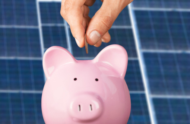 Main déposant une pièce dans une tirelire rose devant des panneaux solaires photovoltaïques pour illustrer les économies d'énergie.