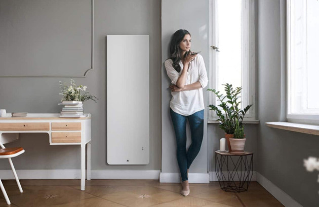Femme appuyée sur le mur à l'intérieur d'une maison à côté d'un radiateur à inertie qui parle au téléphone