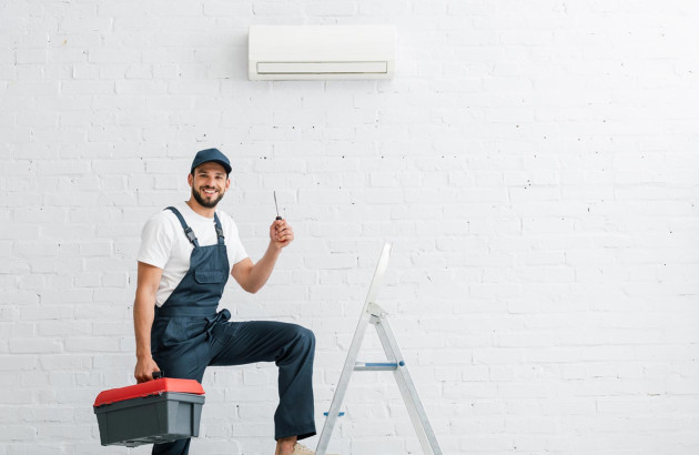 homme souriant en salopette et casquette tenant un tournevis et une boîte à outils et qui monte sur une échelle à côté d'un climatiseur mural blanc