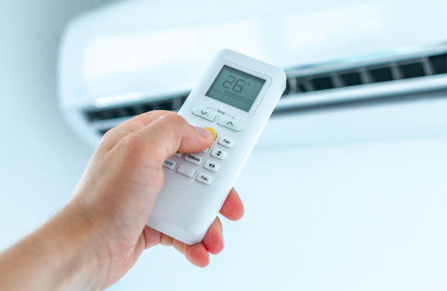 main qui tient une télécommande de climatiseur affichant 26°C avec l'unité de climatisation murale blanche en arrière-plan