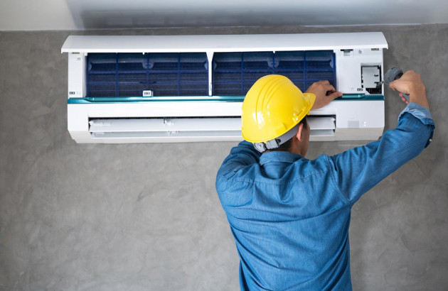 professionnel portant un casque de sécurité jaune et une chemise de travail bleue effectuant la maintenance ou la réparation d'un climatiseur mural blanc