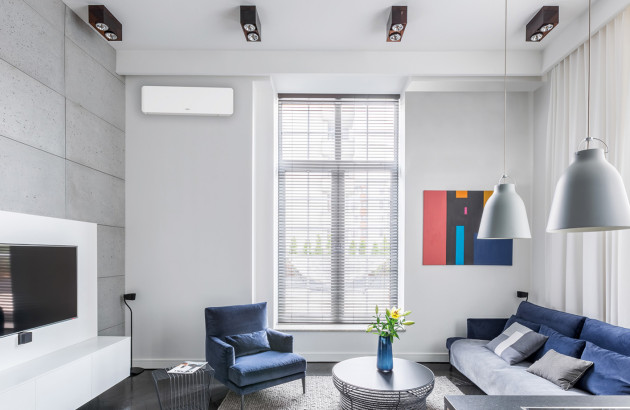 salon moderne et lumineux avec un climatiseur mural blanc avec un grand canapé bleu et un fauteuil et un mur texturé et une fenêtre avec stores