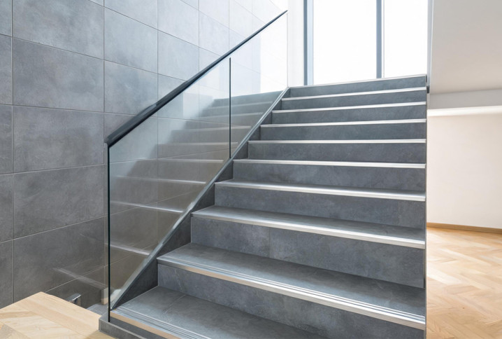 Escalier intérieur en carrelage gris effet béton, avec nez de marche métalliques antidérapants et garde-corps en verre.