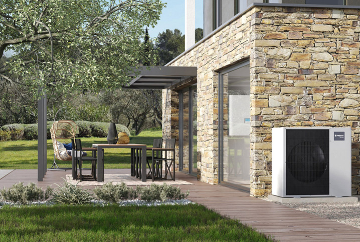 Pompe à chaleur extérieure Frisquet installée contre la façade en pierre d’une maison avec terrasse en bois et pergola.