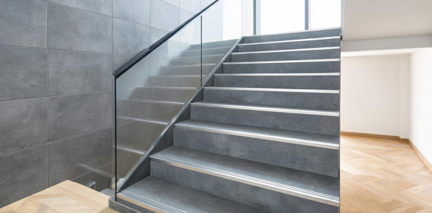 Escalier intérieur en carrelage gris effet béton, avec nez de marche métalliques antidérapants et garde-corps en verre.