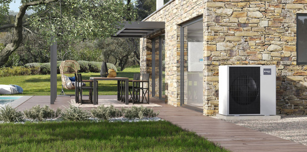 Pompe à chaleur extérieure Frisquet installée contre la façade en pierre d’une maison avec terrasse en bois et pergola.