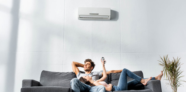 Couple détendu sur un canapé sous un climatiseur mural, contrôlant la température avec une télécommande.
