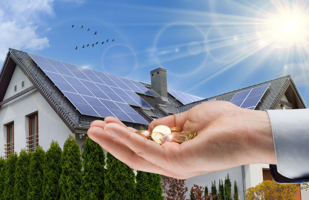 main qui tient des pièces devant une maison équipée de panneaux solaires