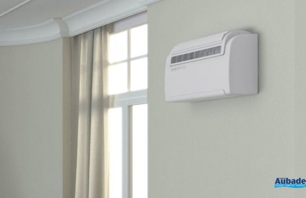Climatiseur monobloc mural blanc installé en hauteur près d’une fenêtre dans un intérieur lumineux.