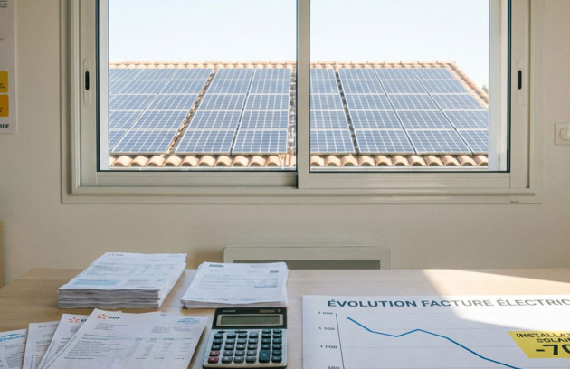 Vue depuis l’intérieur d’une maison sur des panneaux solaires photovoltaïques installés sur la toiture, avec factures d’électricité et calculatrice illustrant l’analyse de la rentabilité solaire.