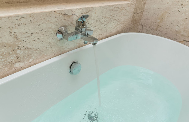 Baignoire remplie d’eau avec mitigeur mural, robinetterie chromée sur carrelage effet pierre.