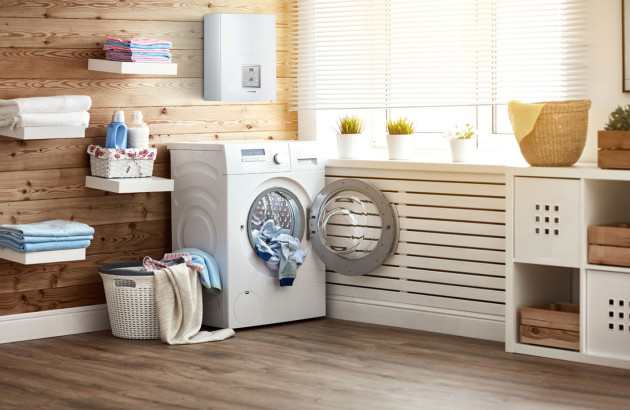 Chaudière électrique murale installée au-dessus d’un lave-linge dans une buanderie moderne avec radiateurs et rangements en bois, illustration d’un système de chauffage central compact et sans fumées.