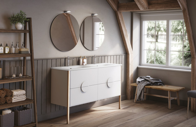 Salle de bain mansardée avec poutres apparentes, meuble double vasque blanc sur pieds fins, deux miroirs ronds et étagère de rangement en bois foncé.